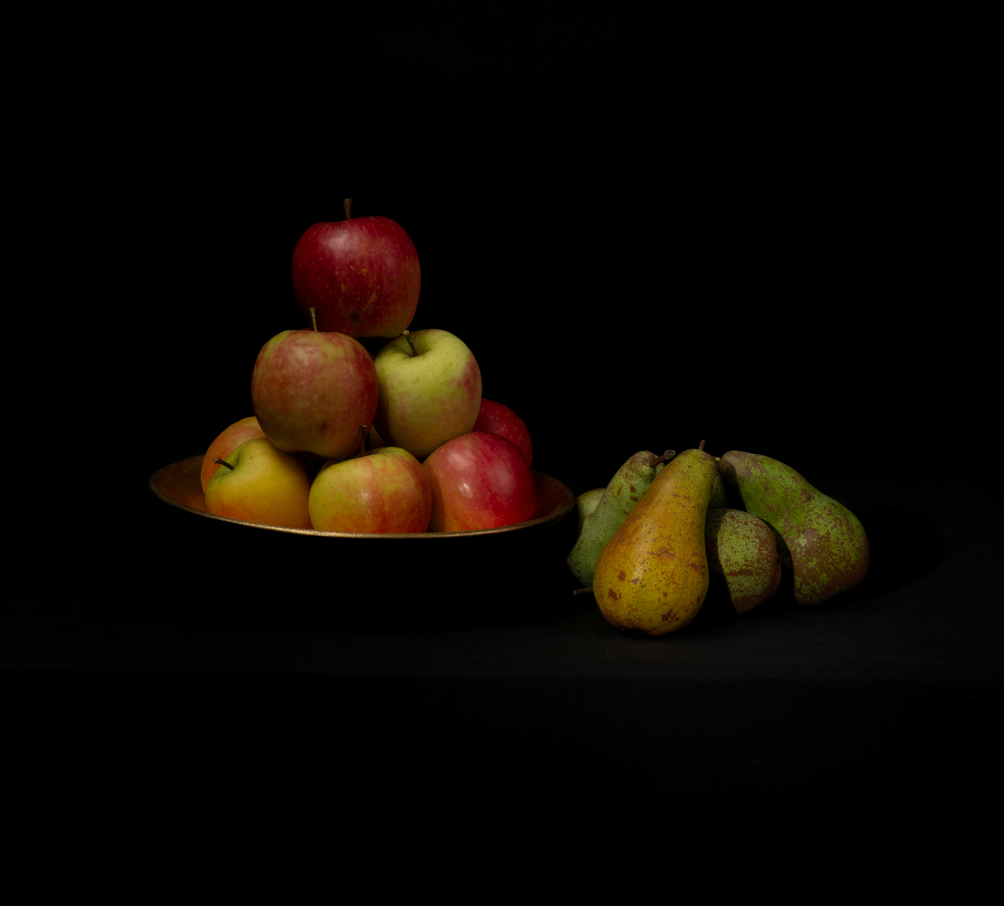 A low-key monochrome still life titled Orderly Apples and Pell-mell Pears. On the left, a group of apples is carefully stacked into a loose pyramid on a shallow dark plate, with a single apple balanced at the top. Their smooth skins and rounded forms are picked out by soft directional light against an almost completely black background. To the right, a contrasting group of pears lies in a more irregular, haphazard heap, their elongated shapes overlapping and their mottled, speckled skins rendered with strong texture. The fruit emerges gradually from deep shadow, while the surrounding surface and background fall almost entirely into black. The arrangement contrasts the formal order of the apples with the casual disorder of the pears. All of the fruit was harvested from the garden.